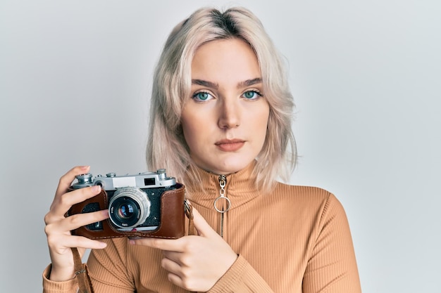 Giovane ragazza bionda che tiene la macchina fotografica vintage rilassata con un'espressione seria sul viso semplice e naturale guardando la telecamera