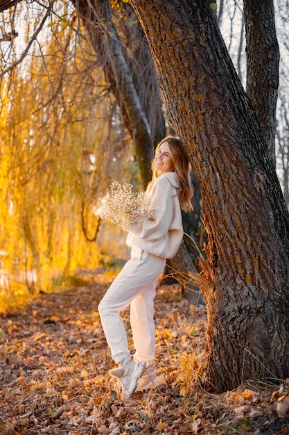 Giovane ragazza bionda con fiori in piedi nel parco autunnale vicino al lago Donna che indossa un maglione beige e pantaloni bianchi Ragazza che posa per una foto in una giornata di sole