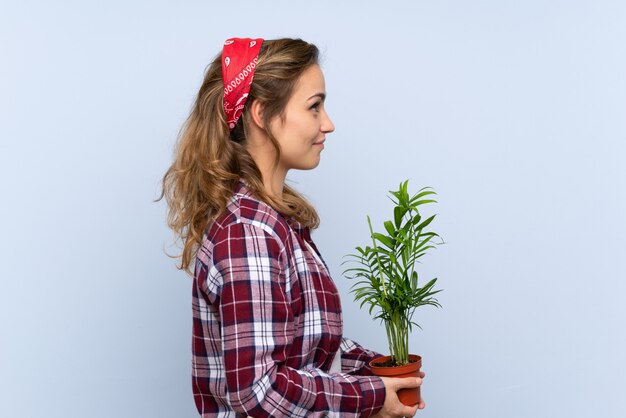 Giovane ragazza bionda del giardiniere che tiene una pianta sopra fondo blu isolato