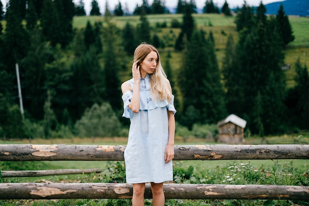 Giovane ragazza bionda elegante attraente in vestito blu che posa vicino al recinto di legno nella campagna