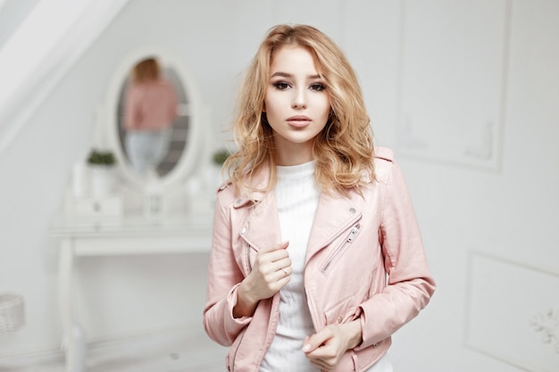 Giovane ragazza bionda in elegante giacca di pelle rosa in una stanza con interni bianchi