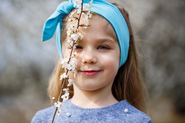 Giovane ragazza bionda in piedi in un giardino fiorito. Ciliegio in fiore. Ritratto di bella bambina. Primo piano del piccolo volto del modello. Sakura in fiore, sera primaverile