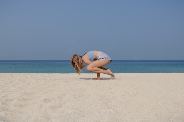 Giovane ragazza bionda pratica yoga e meditazione nella posizione bakasana e posa della gru sulla spiaggia in una giornata di sole.