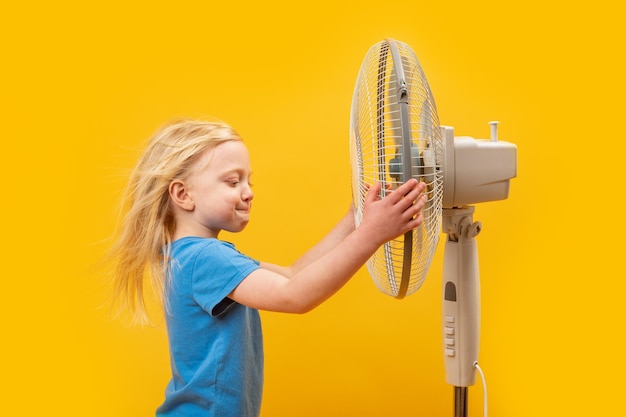 Giovane ragazza bionda premurosa con i capelli mossi dal vento vicino al ventilatore sfondo giallo Bambino Fairhaired vicino al ventilatore quando fa caldo