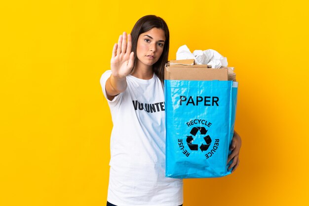 Giovane ragazza brasiliana che tiene un sacchetto di riciclaggio pieno di carta da riciclare isolato su priorità bassa gialla che fa gesto di arresto