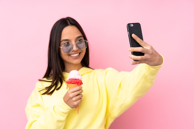 Giovane ragazza bruna che tiene un gelato alla cornetta su sfondo rosa isolato facendo un selfie