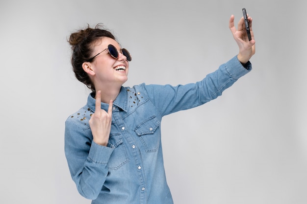 Giovane ragazza bruna in occhiali rotondi. I peli sono raccolti in una crocchia. Ragazza con un telefono nero. La ragazza fa selfie.