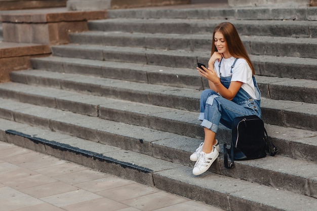 Giovane ragazza carina con la cartella seduta sulle scale e la scrittura di sms sul telefono