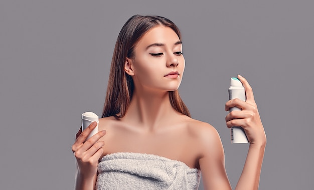 Giovane ragazza castana attraente con capelli sciolti con deodorante stick e aerosol antitraspirante isolato su sfondo grigio. Cura del corpo.