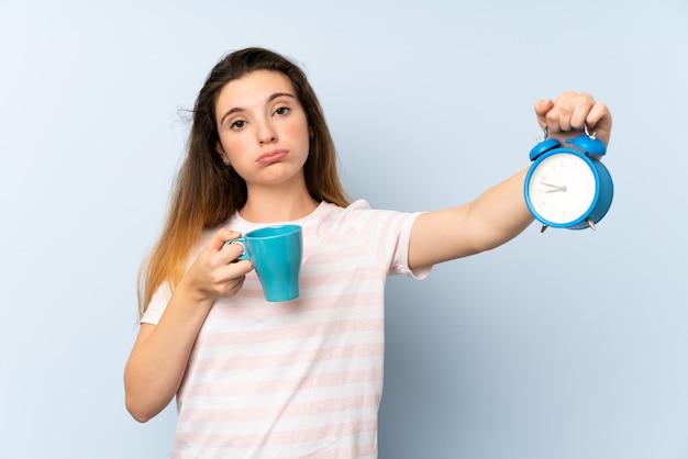 Giovane ragazza castana che tiene una tazza di caffè e un orologio d'annata sopra la parete isolata