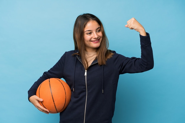 Giovane ragazza castana sopra il blu isolato con la palla di pallacanestro