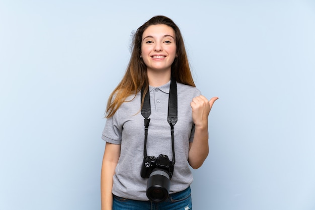 Giovane ragazza castana sopra la parete blu isolata con una macchina fotografica professionale e che indica il lato