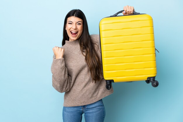 Giovane ragazza castana sopra la parete blu isolata in vacanza con la valigia di viaggio