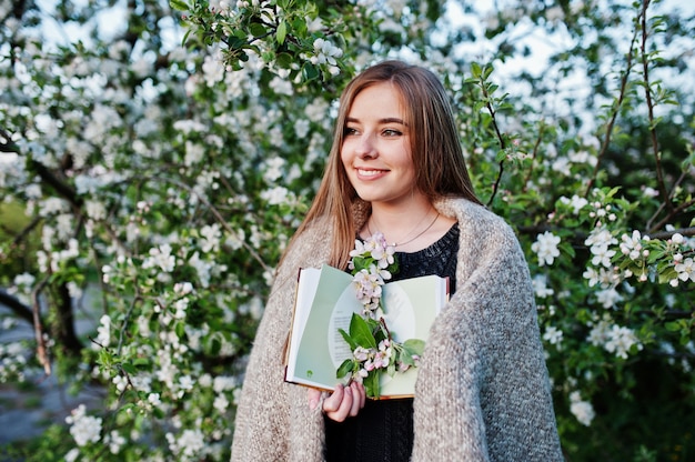 Giovane ragazza castana sul plaid contro l'albero del fiore della molla e tenere il libro a portata di mano.