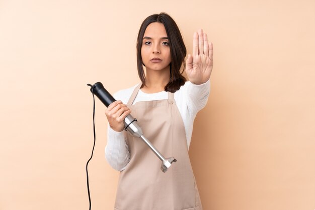 Giovane ragazza castana usando il frullatore a immersione isolato