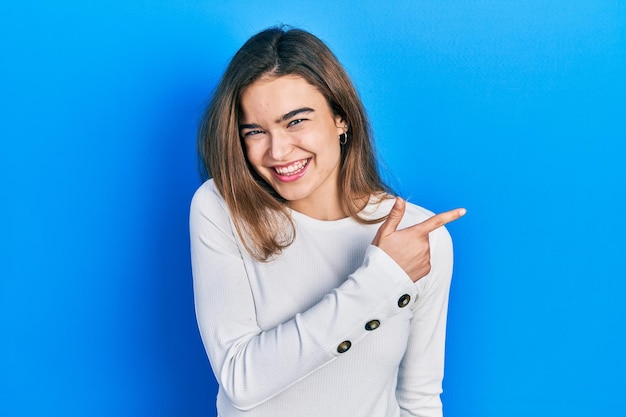 Giovane ragazza caucasica che indossa abiti casual sorridendo allegro indicando con la mano e il dito fino al lato