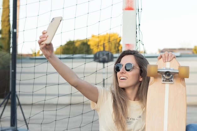 Giovane ragazza caucasica che ride mentre si fa un selfie seduto accanto a una rete con il suo longboard