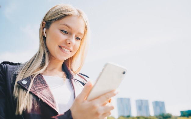 Giovane ragazza caucasica con cuffie wireless nel parco utilizzando il telefono tablet e sorridente