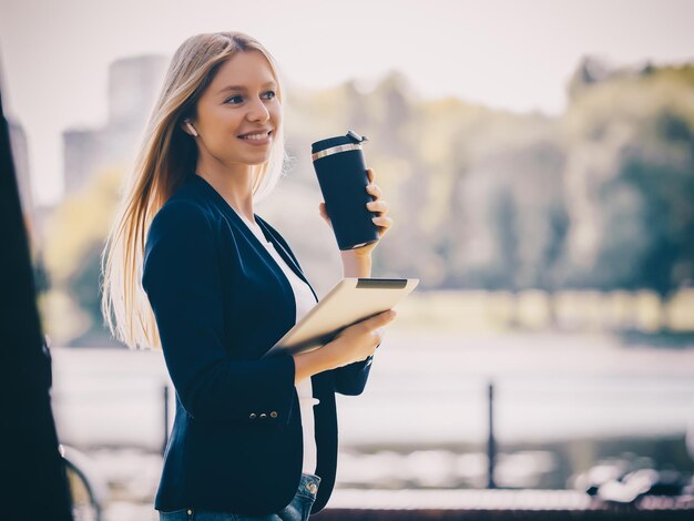 Giovane ragazza caucasica con cuffie wireless nel parco utilizzando il telefono tablet e sorridente