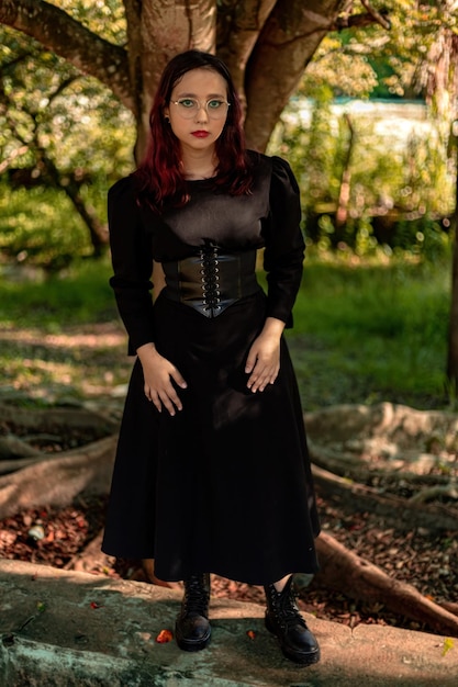 Giovane ragazza caucasica dai capelli scuri con striature rosse in abito nero con cintura a corsetto e occhiali in posa per un servizio di bellezza femminile