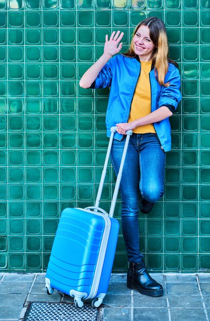 giovane ragazza caucasica salutando un'altra persona appoggiata a un muro verde con una valigia di rotolamento blu