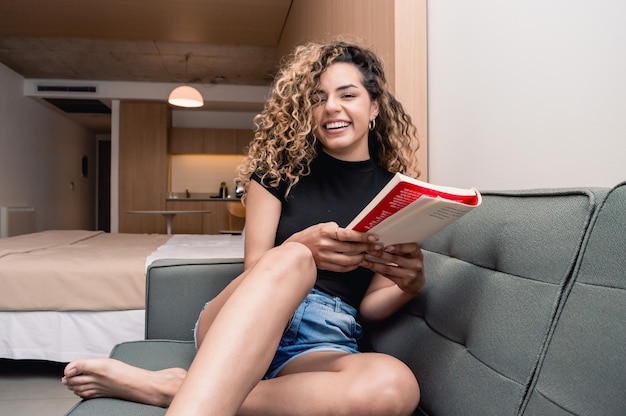 Giovane ragazza caucasica seduta sul divano nel suo appartamento con la cucina e il suo letto dietro di lei con in mano un libro che sorride felicemente guardando la telecamera