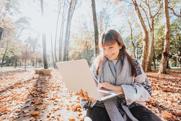 Giovane ragazza caucasica streamer fuori usando un laptop per controllare il lavoro, atteggiamento felice e sorridente. Stile giovane e moderno alla moda. Stagione autunnale. Lavorare ovunque, libertà finanziaria, investimenti