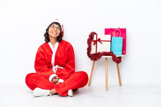 Giovane ragazza che celebra il Natale seduta sul pavimento isolato su sfondo bianco alzando lo sguardo mentre sorride