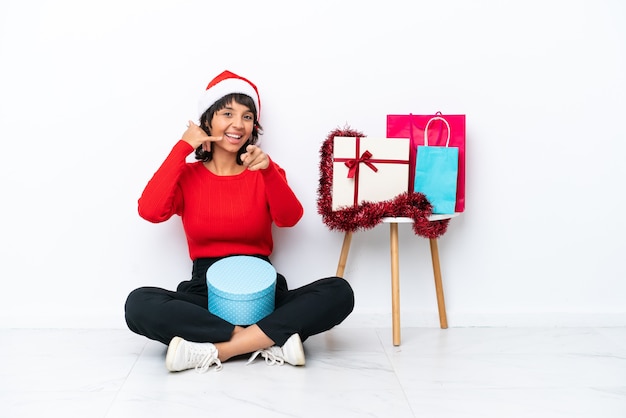 Giovane ragazza che celebra il Natale seduta sul pavimento isolato su sfondo bianco che fa il gesto del telefono e indica la parte anteriore