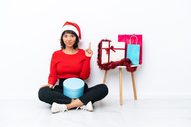 Giovane ragazza che celebra il Natale seduta sul pavimento isolato su sfondo bianco che indica con il dito indice una grande idea