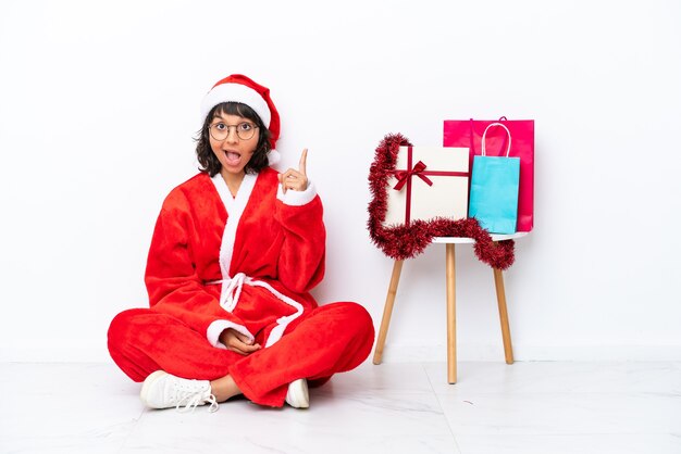 Giovane ragazza che celebra il Natale seduta sul pavimento isolato su sfondo bianco che indica con il dito indice una grande idea