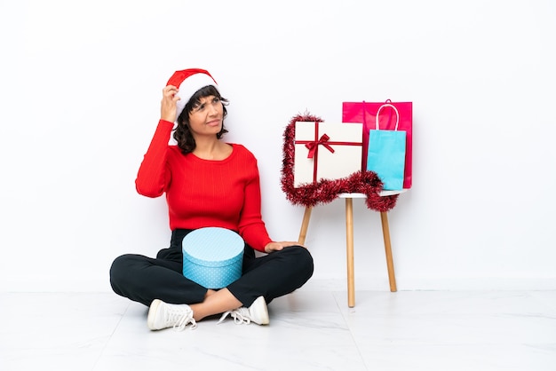 Giovane ragazza che celebra il Natale seduta sul pavimento isolato su sfondo bianco con dubbi e con un'espressione del viso confusa