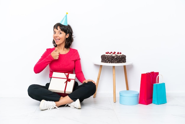 Giovane ragazza che celebra il suo compleanno seduto sul pavimento isolato su sfondo bianco dando un pollice in alto gesto