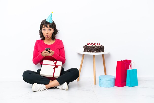 Giovane ragazza che celebra il suo compleanno seduto sul pavimento isolato su sfondo bianco sorpreso e inviando un messaggio