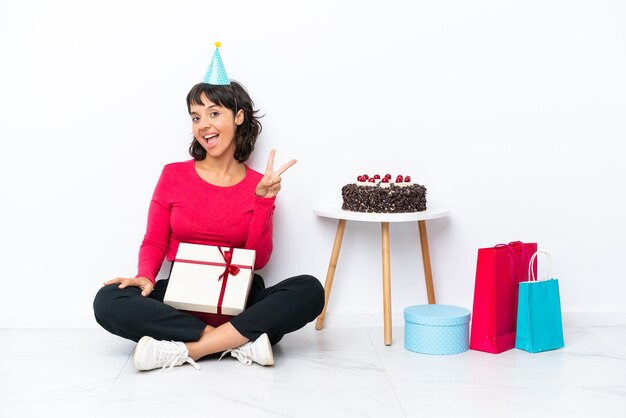 Giovane ragazza che festeggia il suo compleanno seduta sul pavimento isolato su sfondo bianco sorridendo e mostrando il segno della vittoria