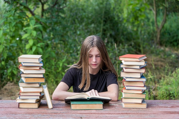 Giovane ragazza che legge un libro in giardino a un tavolo di legno con una pila di libri