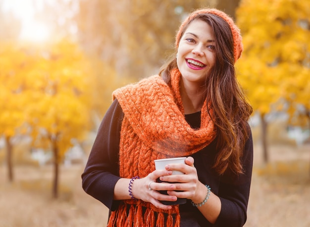 Giovane ragazza che ride con una tazza di caffè (tè) per una passeggiata nel parco all'aperto. Tempo autunnale. I raggi del sole illuminano il modello da dietro.