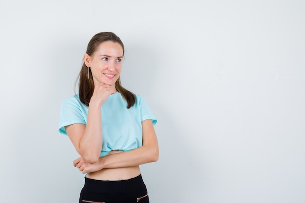 Giovane ragazza che si appoggia il mento a portata di mano, in piedi di lato in maglietta turchese, pantaloni e sembra curiosa.