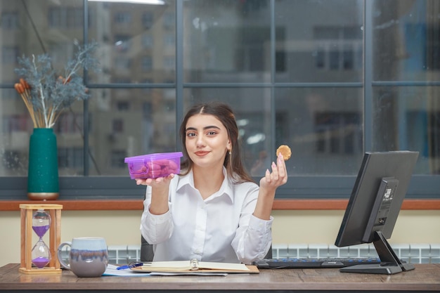 Giovane ragazza che tiene un contenitore per biscotti e un biscotto mentre era seduto in ufficio