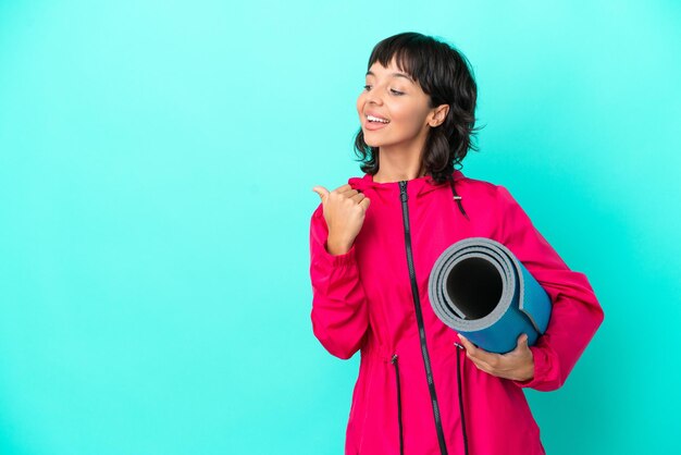 Giovane ragazza che va a lezioni di yoga mentre tiene un tappetino isolato su sfondo blu che punta al lato per presentare un prodotto