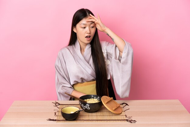 Giovane ragazza cinese che indossa il kimono e mangia le tagliatelle facendo il gesto di sorpresa mentre guarda al lato