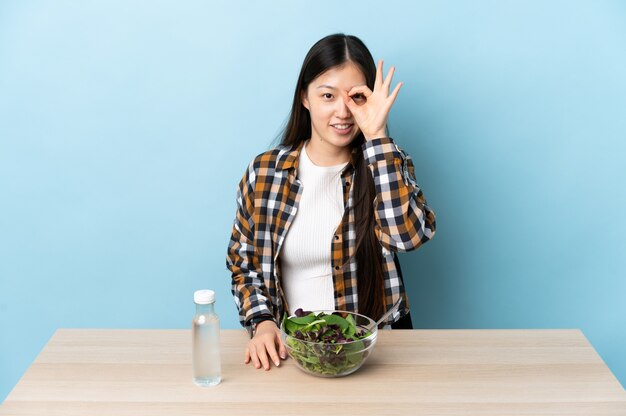 Giovane ragazza cinese che mangia un'insalata che mostra segno giusto con le dita