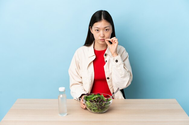 Giovane ragazza cinese che mangia un'insalata che mostra un segno del gesto di silenzio