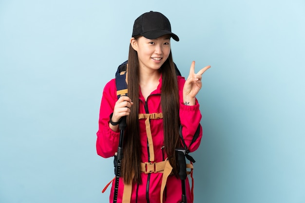 Giovane ragazza cinese con zaino e bastoncini da trekking sopra il blu isolato che sorride e che mostra il segno di vittoria