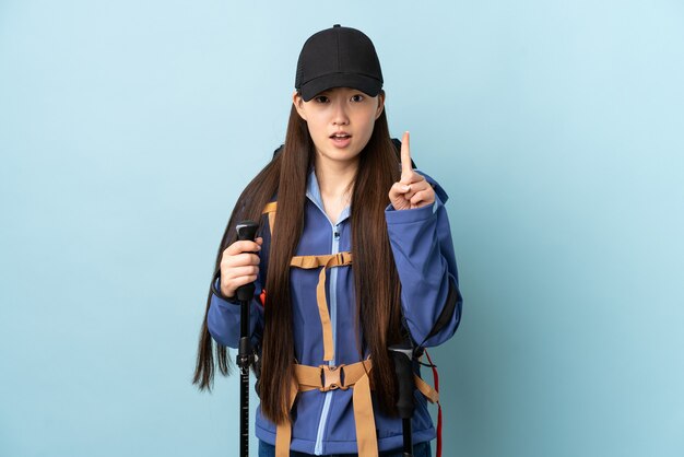 Giovane ragazza cinese con zaino e bastoncini da trekking su blu contando uno con espressione seria