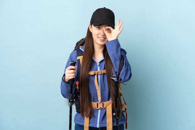 Giovane ragazza cinese con zaino e bastoncini da trekking sul blu isolato che mostra segno giusto con le dita