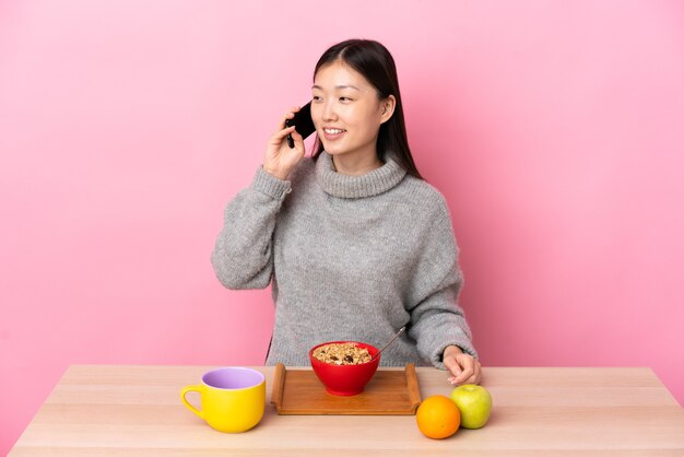 Giovane ragazza cinese facendo colazione in un tavolo mantenendo una conversazione con il telefono cellulare con qualcuno