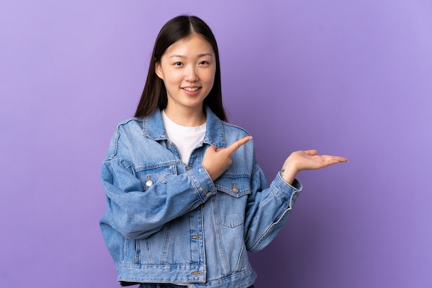 Giovane ragazza cinese sopra la parete viola isolata che tiene il copyspace immaginario sul palmo per inserire un annuncio