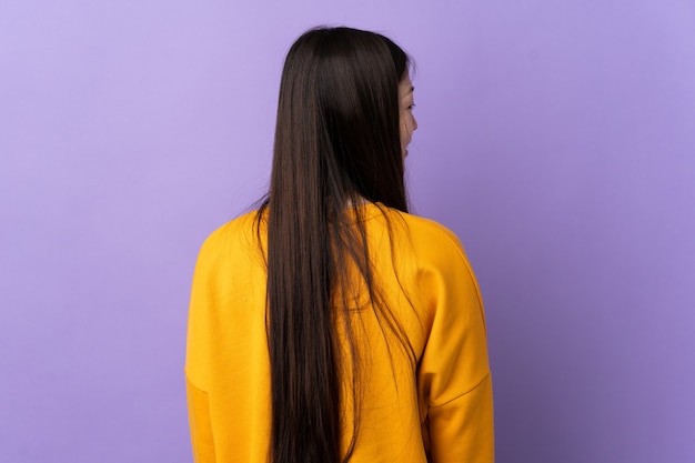 Giovane ragazza cinese su sfondo viola isolato in posizione posteriore e guardando il lato