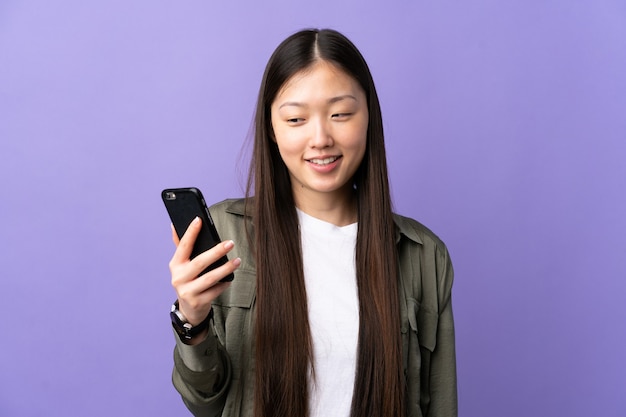 Giovane ragazza cinese utilizzando il telefono cellulare sopra la parete viola isolata con l'espressione felice